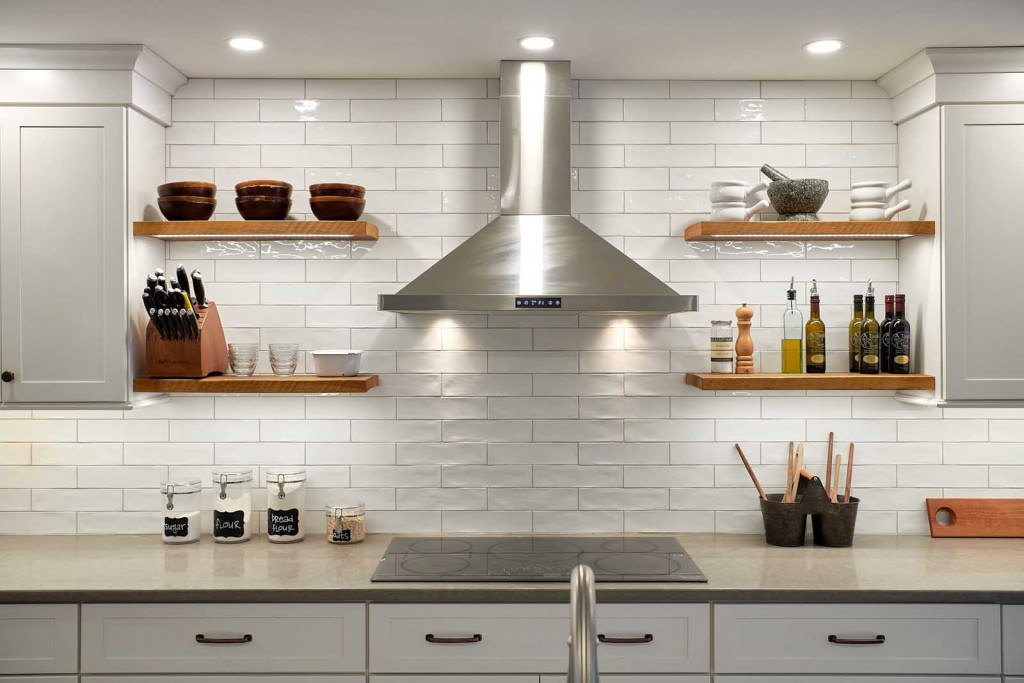 Exton Nordic white kitchen range wall with stainless chimney hood, reclaimed-wood floating shelves, and elongated subway tile