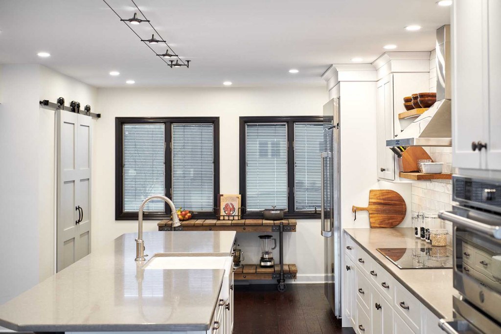 Exton kitchen long view showing white Shaker frameless cabinets, island with fireclay farmhouse sink, black-trimmed windows, and sliding barn door