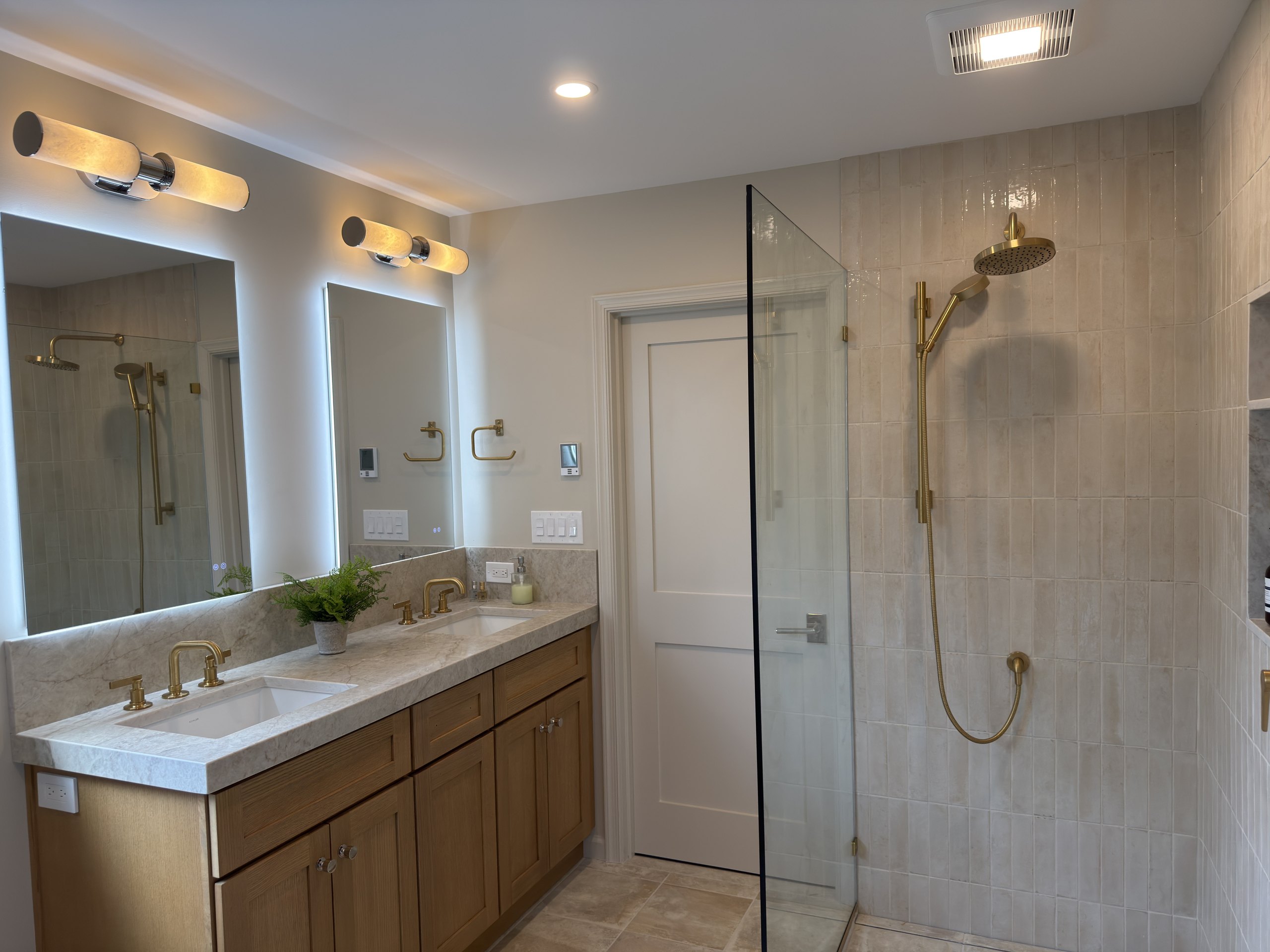 Rift-cut white oak double vanity with two back-lit mirrors, brass sconces, and a frameless glass shower beyond a hinged door