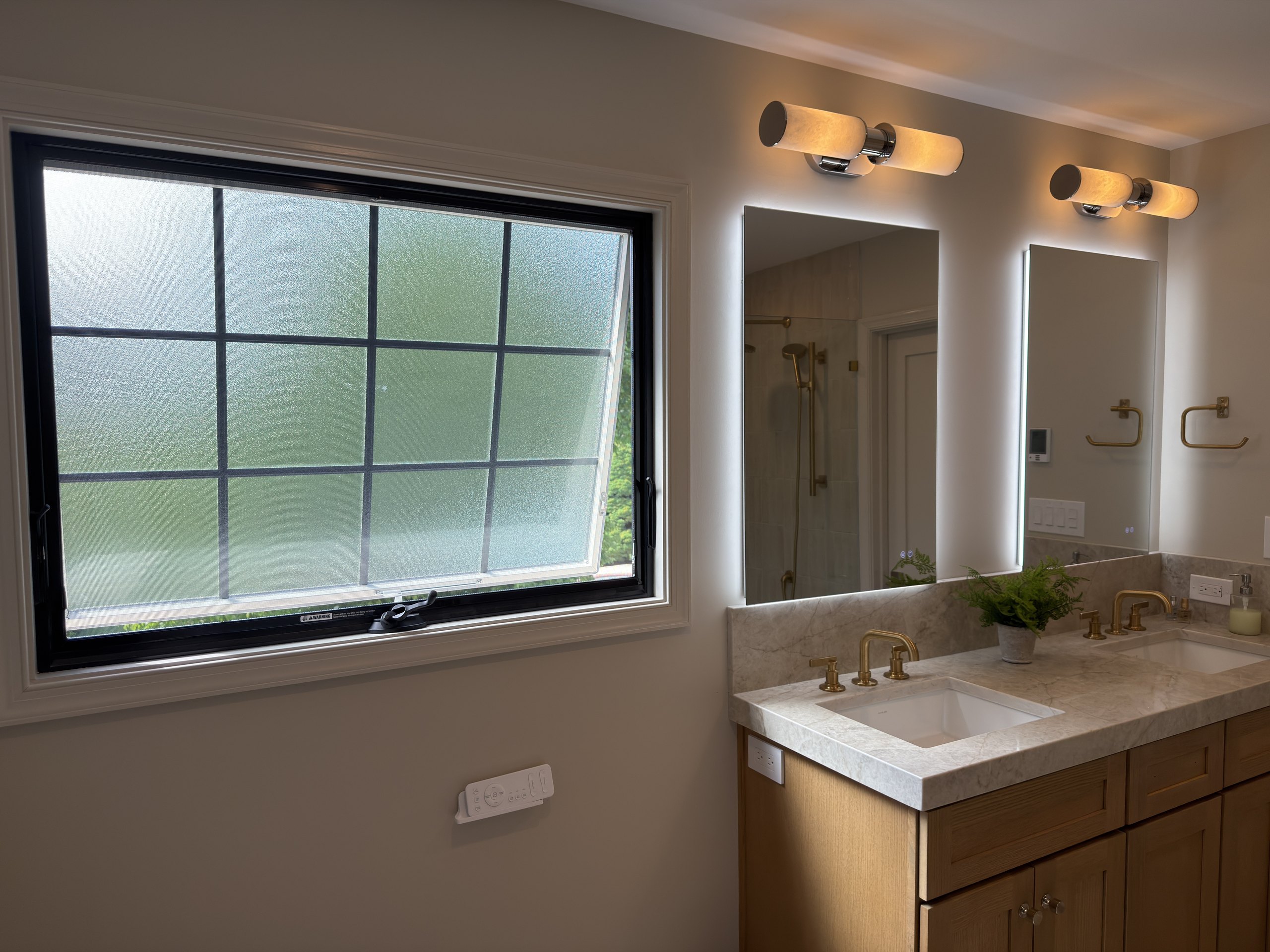 New Andersen awning window with privacy glass above the double vanity, back-lit mirrors, and brass faucets