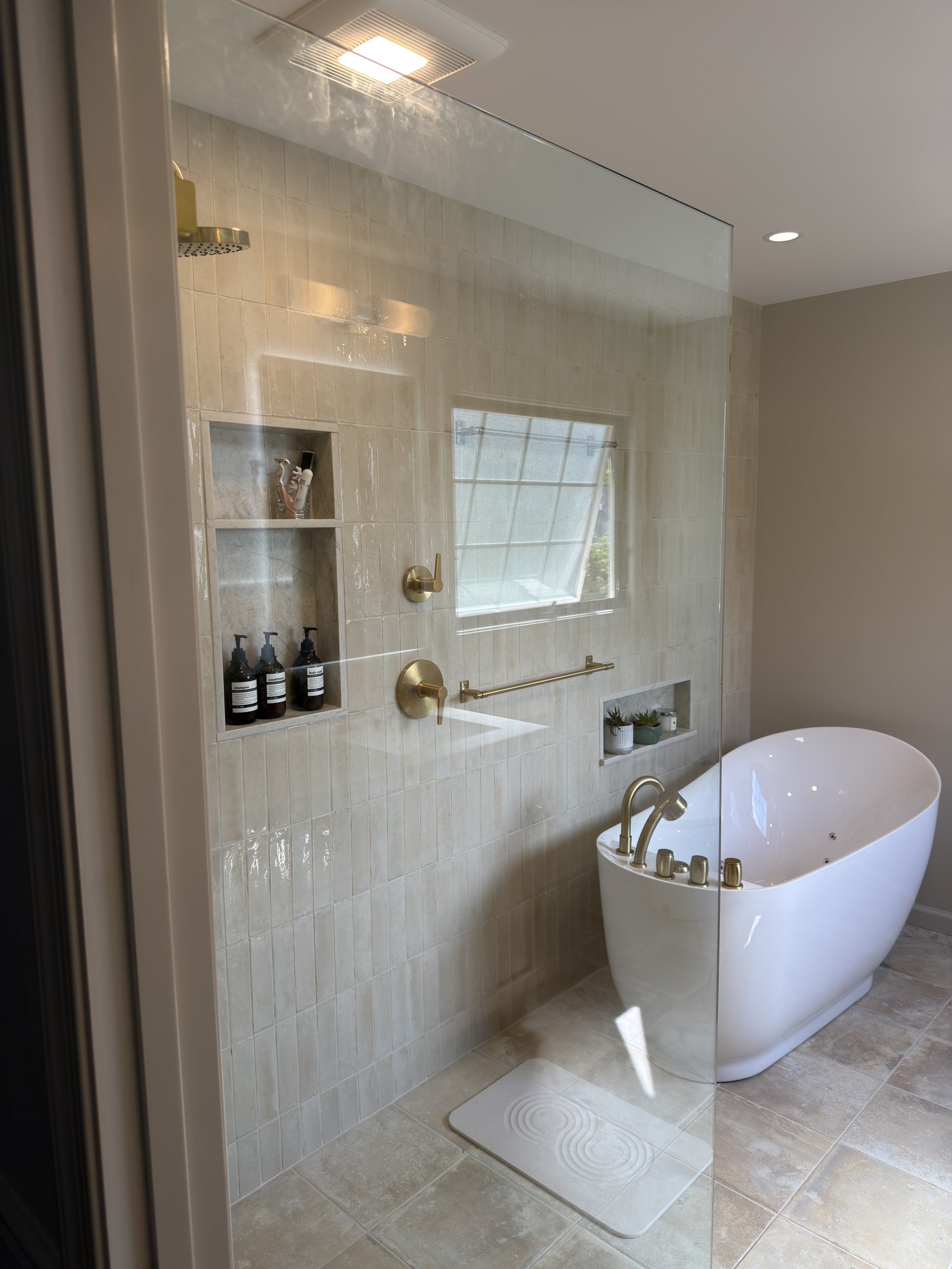 Walk-in shower with brass shower head, quartz-framed niche, and a privacy window beside the freestanding soaking tub
